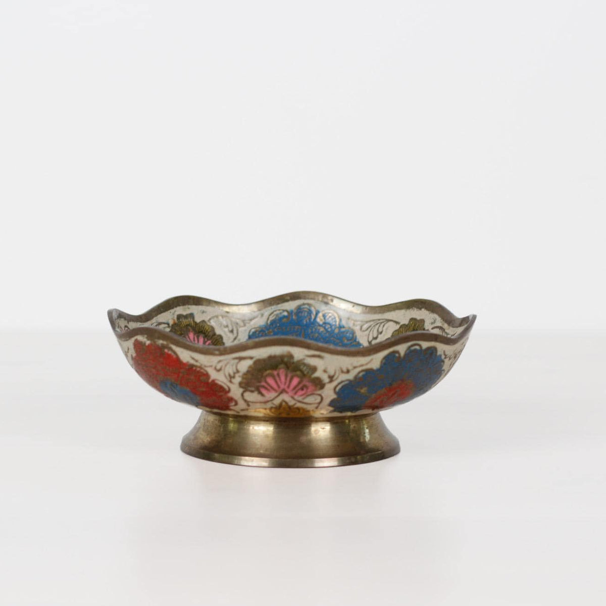A small decorative metal bowl with a scalloped rim, featuring hand-painted floral and mountain motifs in red, blue, pink, and green on a gold-toned base, set against a plain white background.