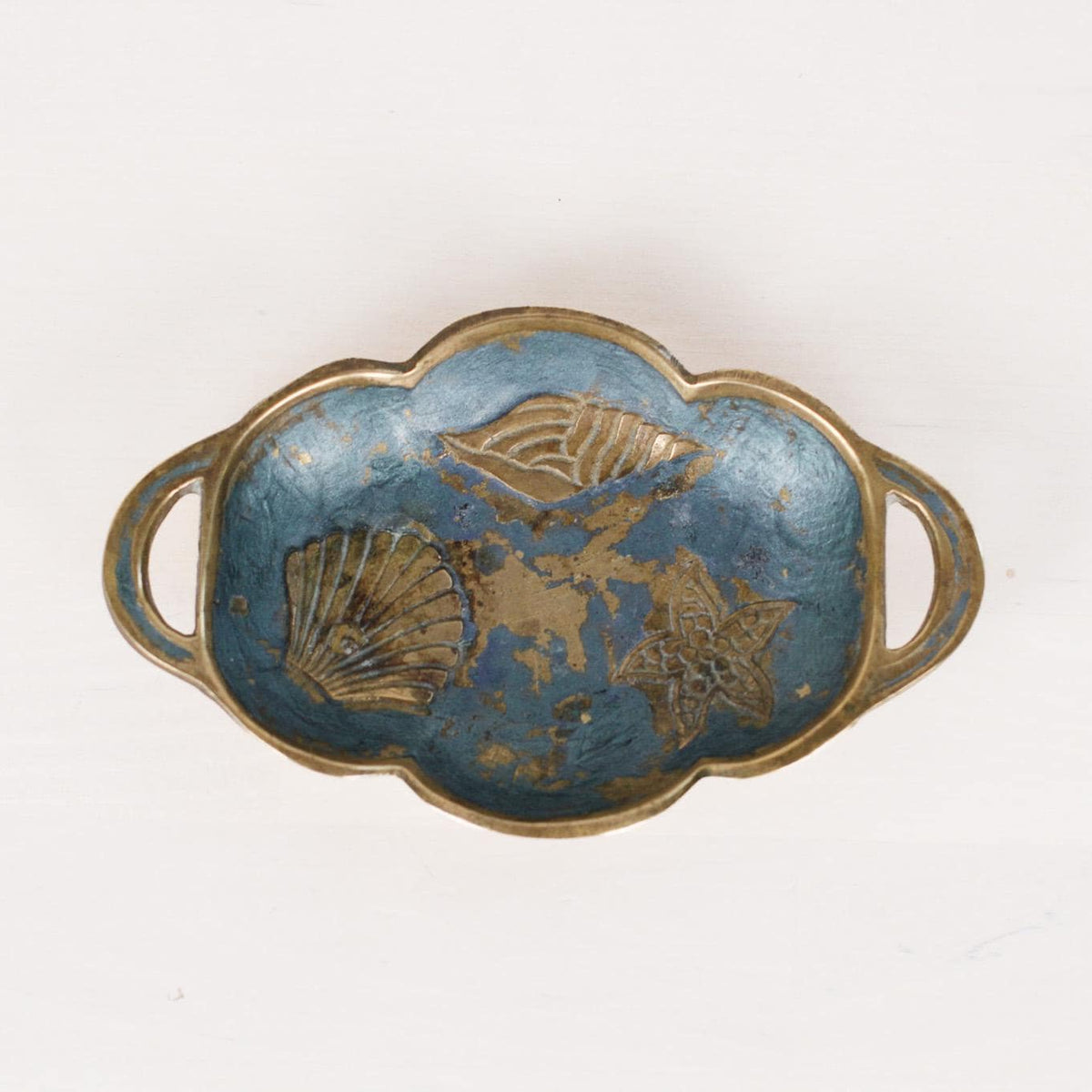 An oval-shaped decorative tray with scalloped edges and loop handles, featuring a blue enamel-like center with gold-toned floral or leaf motifs and a gold-toned rim, shown on a plain white background.