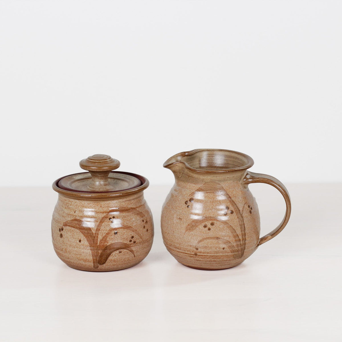 A pair of handmade stoneware ceramic items, including a small lidded jar and a small pitcher, both decorated with a brown floral pattern on a warm beige glazed base, set against a plain white background.