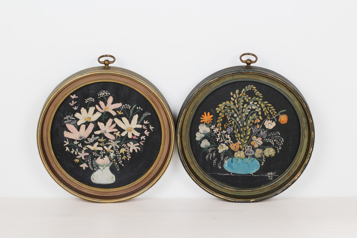 Two round framed floral wall hangings side by side on a white background. The left one has a gold-toned frame and a black background with pink and white flowers in a vase.