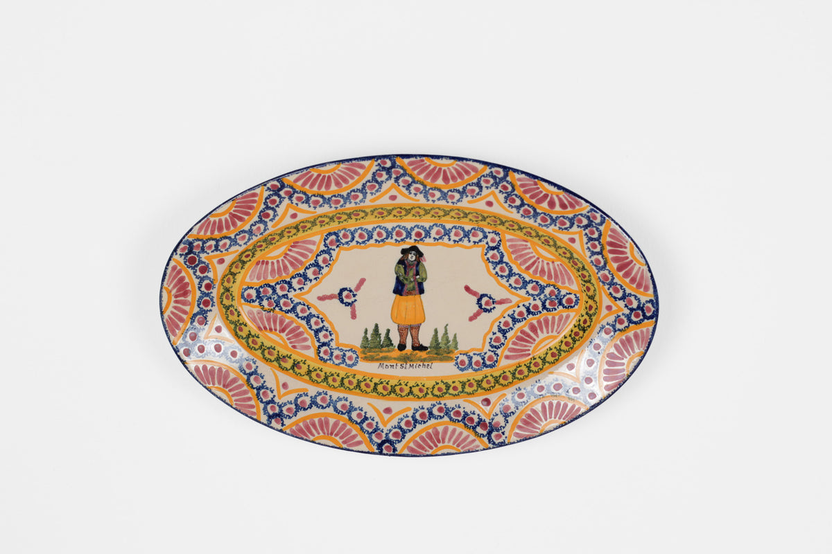 An oval ceramic plate with a colorful hand-painted folk art design, featuring a central figure in a yellow skirt and a decorative border with blue, orange, and yellow patterns, set against a plain light gray background.