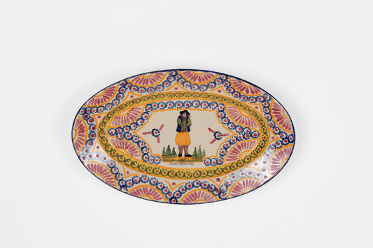 An oval ceramic plate with a colorful hand-painted folk art design, featuring a central figure in a yellow skirt and a decorative border with blue, orange, and yellow patterns, set against a plain light gray background.