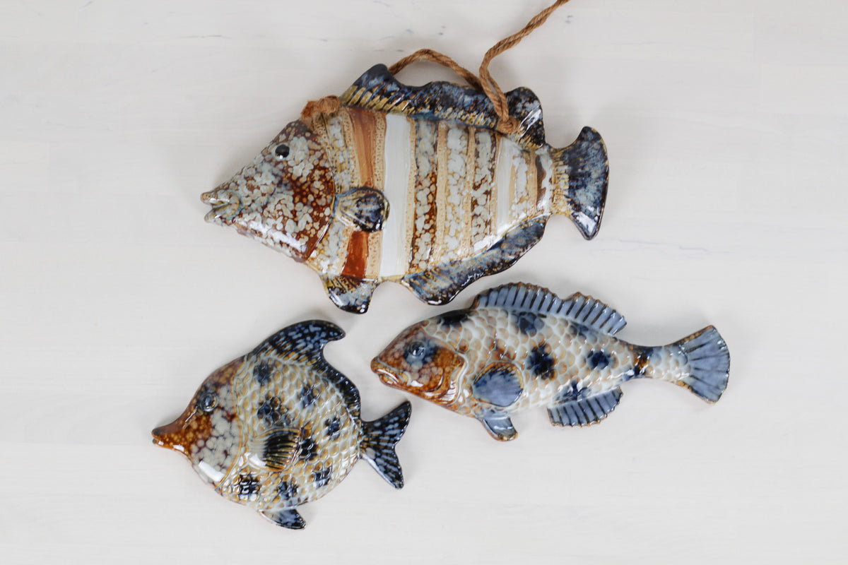 Three decorative fish-shaped ornaments with a glossy finish, featuring mottled patterns of blue, brown, beige, and white, displayed on a light off-white background.