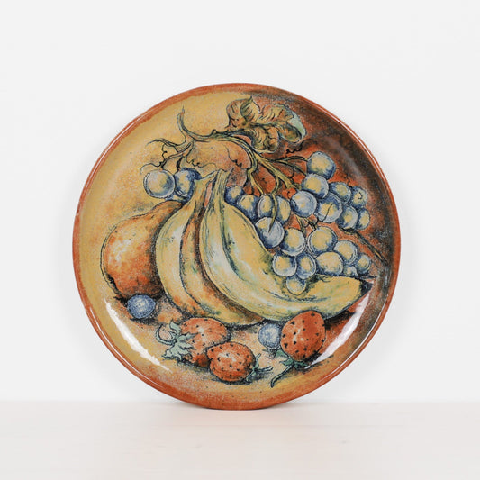 A round ceramic plate featuring a colorful painted still life of fruit, including bananas, grapes, strawberries, and pears, set against a plain white background.