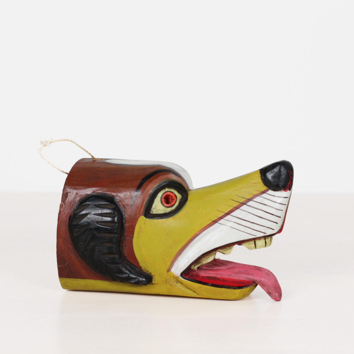 A hand-painted wooden dog head hanging ornament with a white string loop, featuring a yellow-green face, brown head, black ears, and an open mouth showing a pink tongue and white teeth.