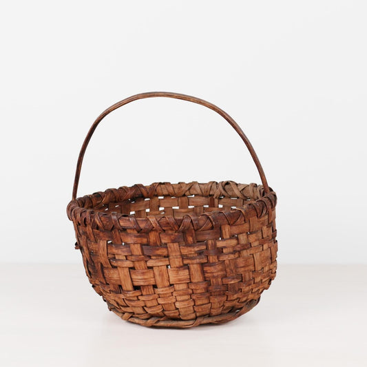 A small hand-woven basket with a single arched handle, made from brown woven strips, set against a plain white background.